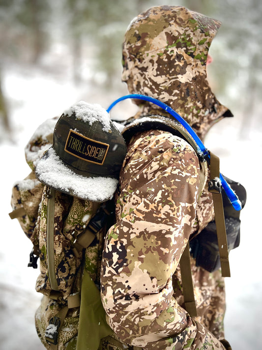 A deep Eddy SnapBack dark multicam hat is attached to a hunting pack, covered in snow, on the back of a hunter wearing camo