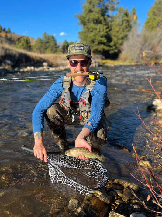 A fly fisherman wearing a West Desert light multicam hat, holding a small fish with fly rod in his mouth celebrating a catch