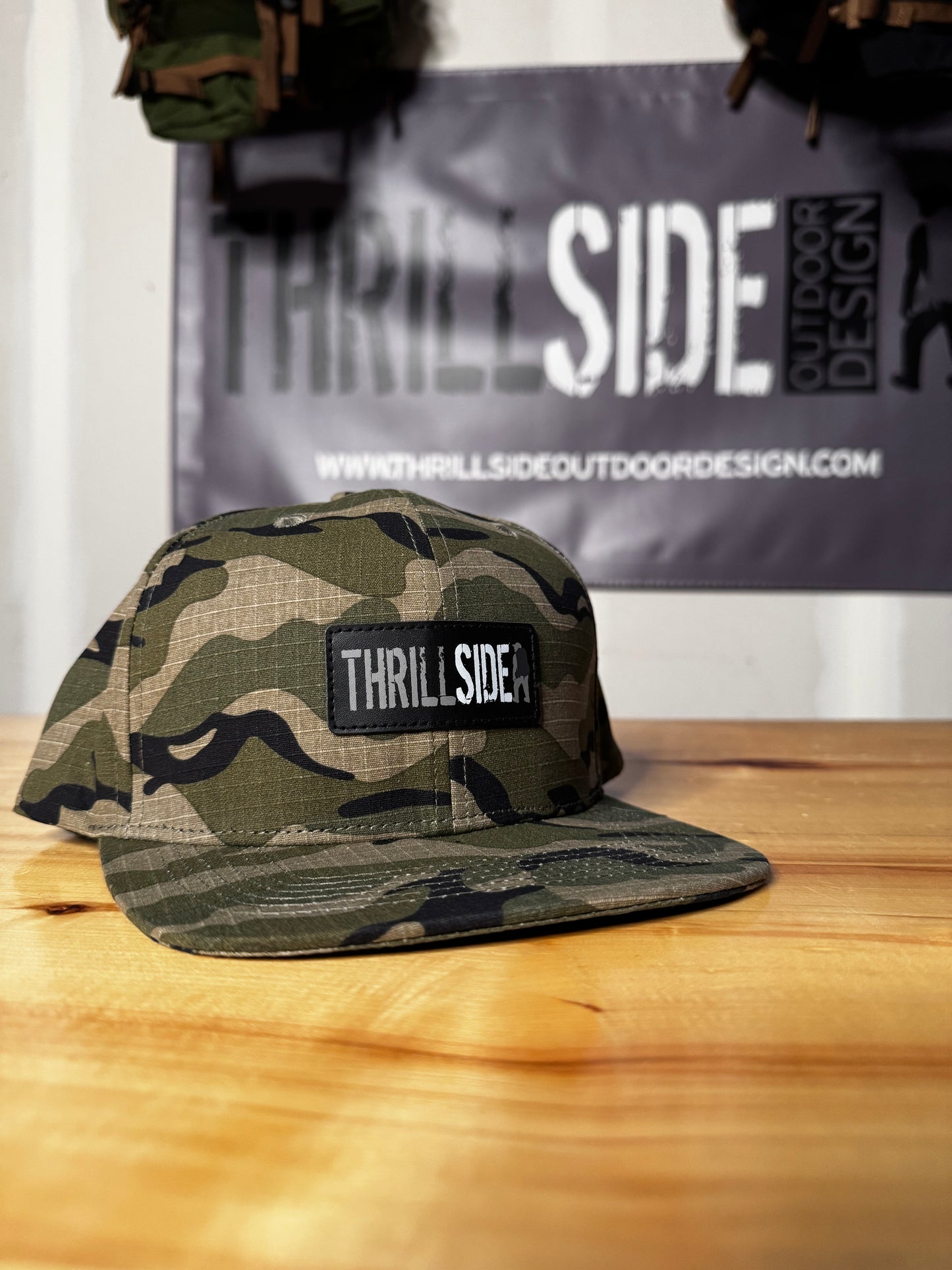 A west desert light multicam hat placed on a wooden table for display, Thrillside banner in the background